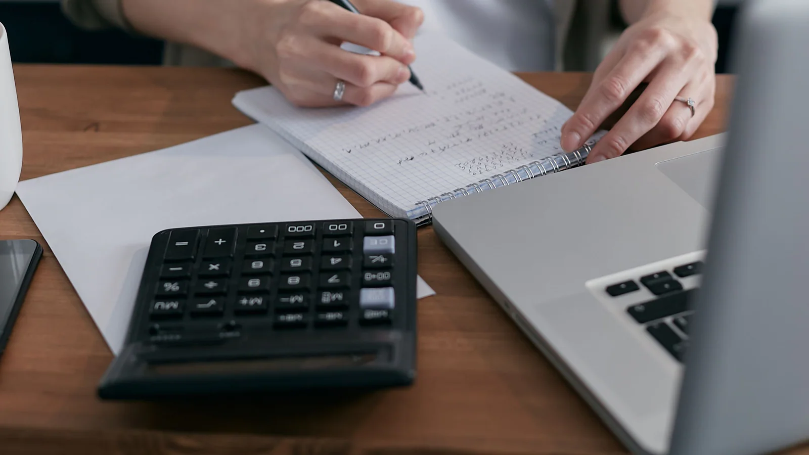 Adult writing calculations in a notebook beside a calculator and laptop
