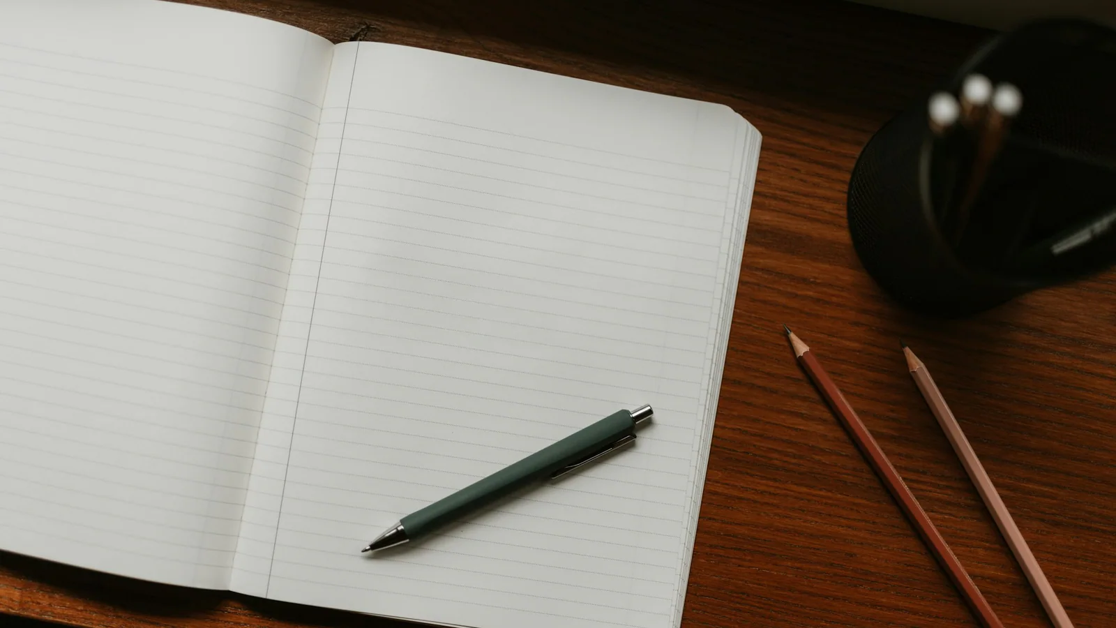 Blank lined notebook with a pen and coloured pencils on a wooden desk