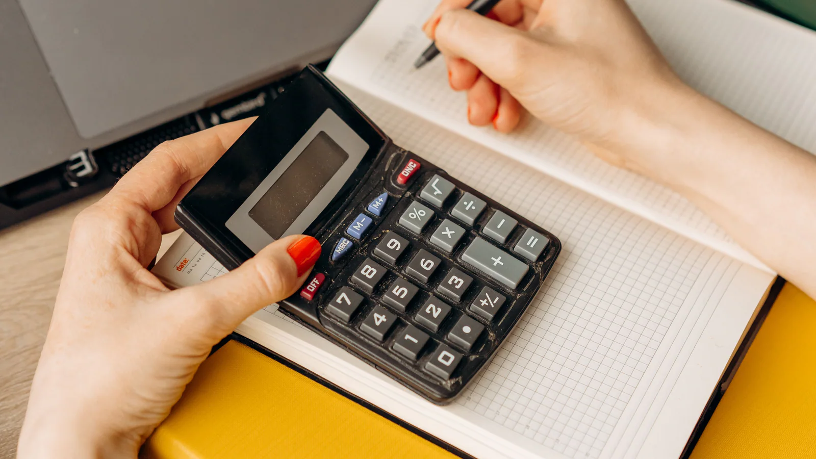Hands using a calculator beside a blank notebook and laptop