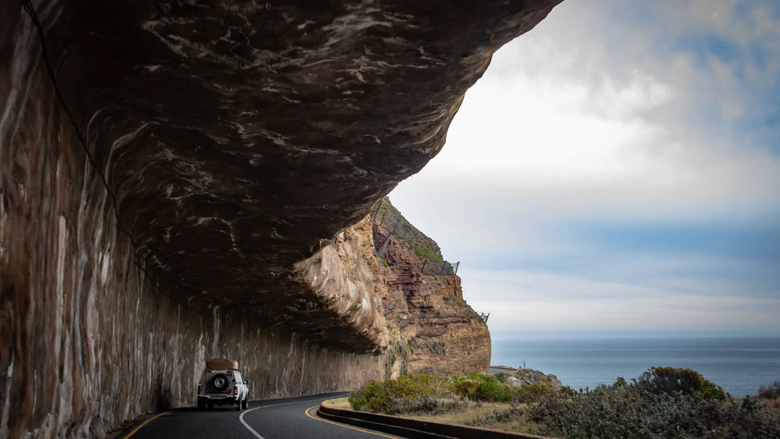 Vehicle driving beneath a rock overhang on a coastal road in Cape Town