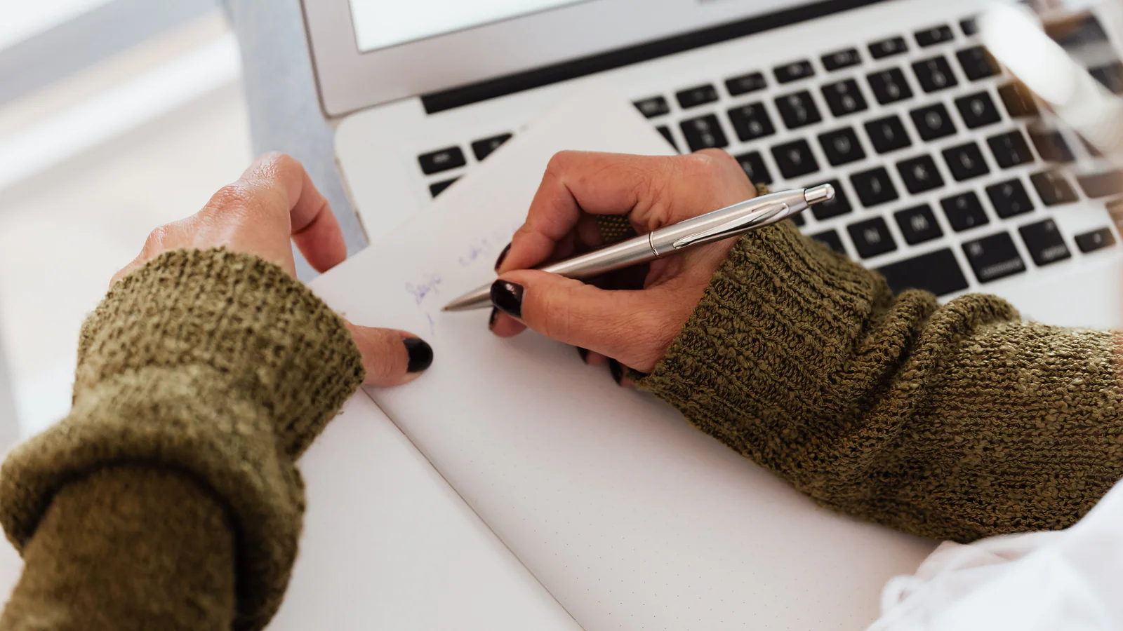 Person taking handwritten notes beside an open laptop