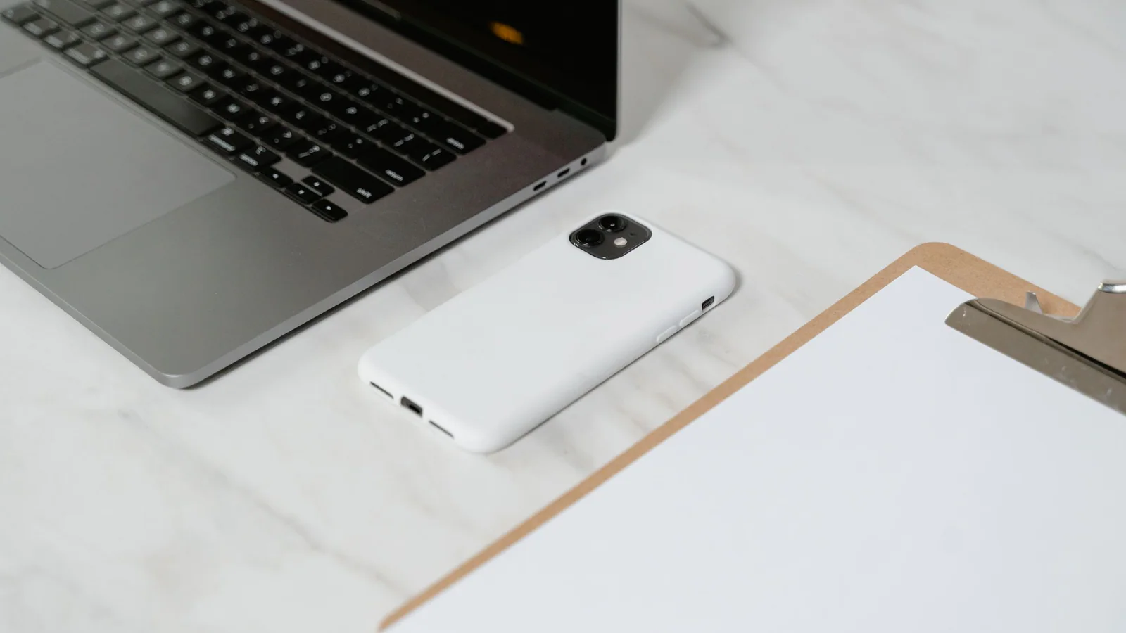 Blank clipboard beside a smartphone and laptop on a white desk