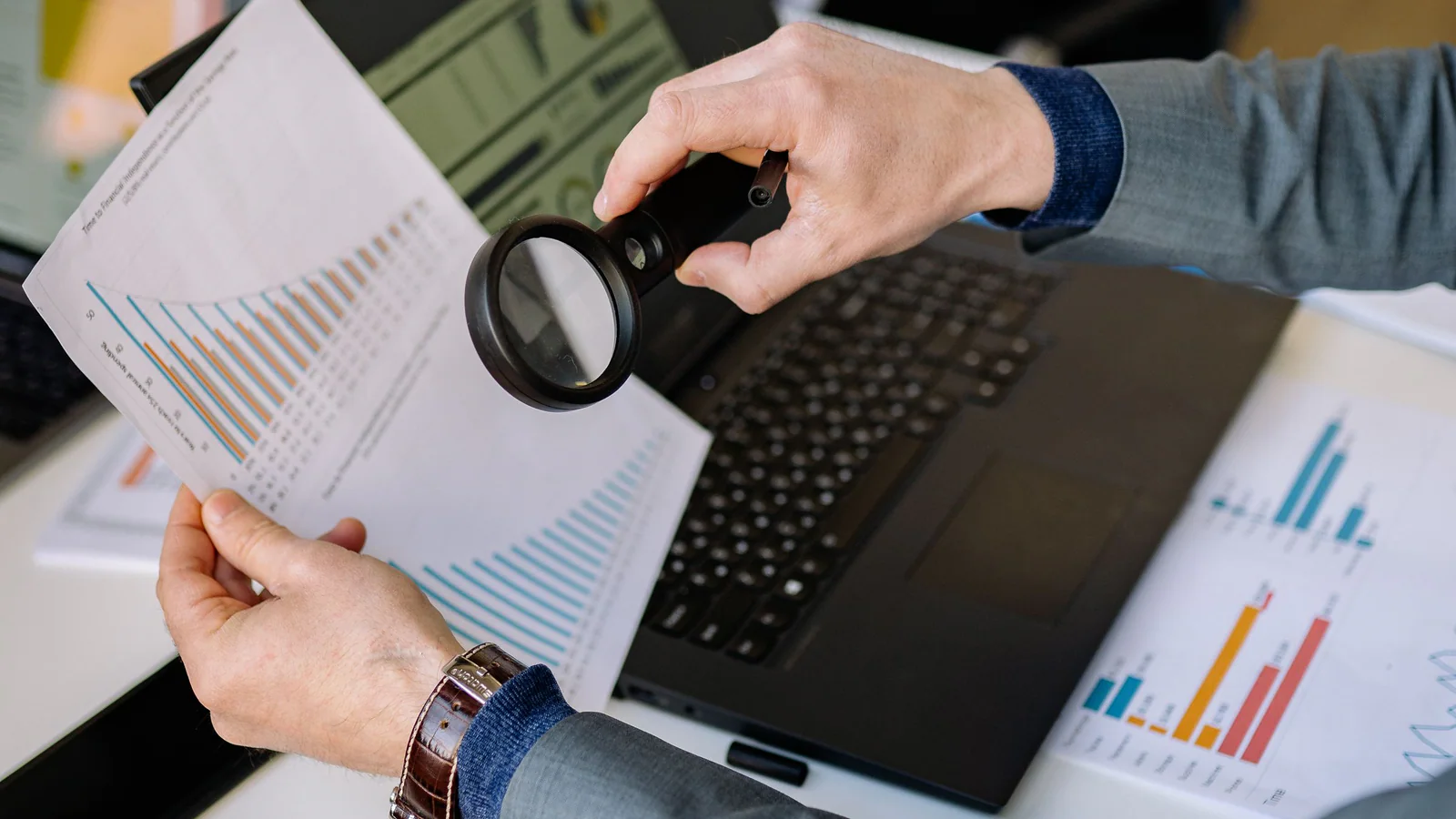 Hands holding a magnifying glass over printed charts at a desk