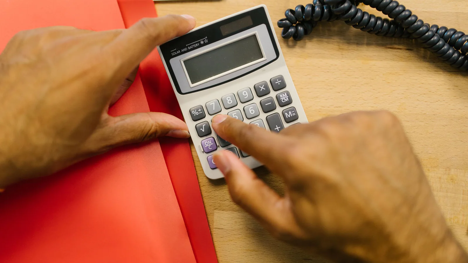 Adult hands using a calculator on a wooden desk beside plain red folders