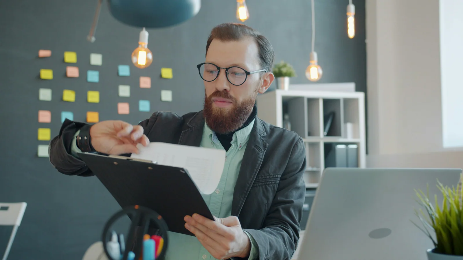 Person wearing glasses reviewing a clipboard beside a laptop in an office
